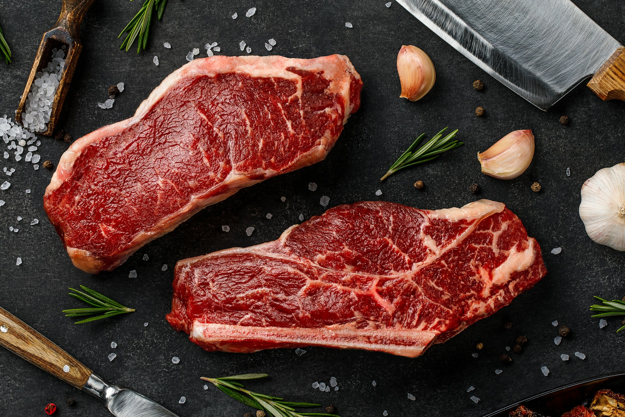 Raw striploin and bone-steak with knife, rosemary, salt and pepper top view